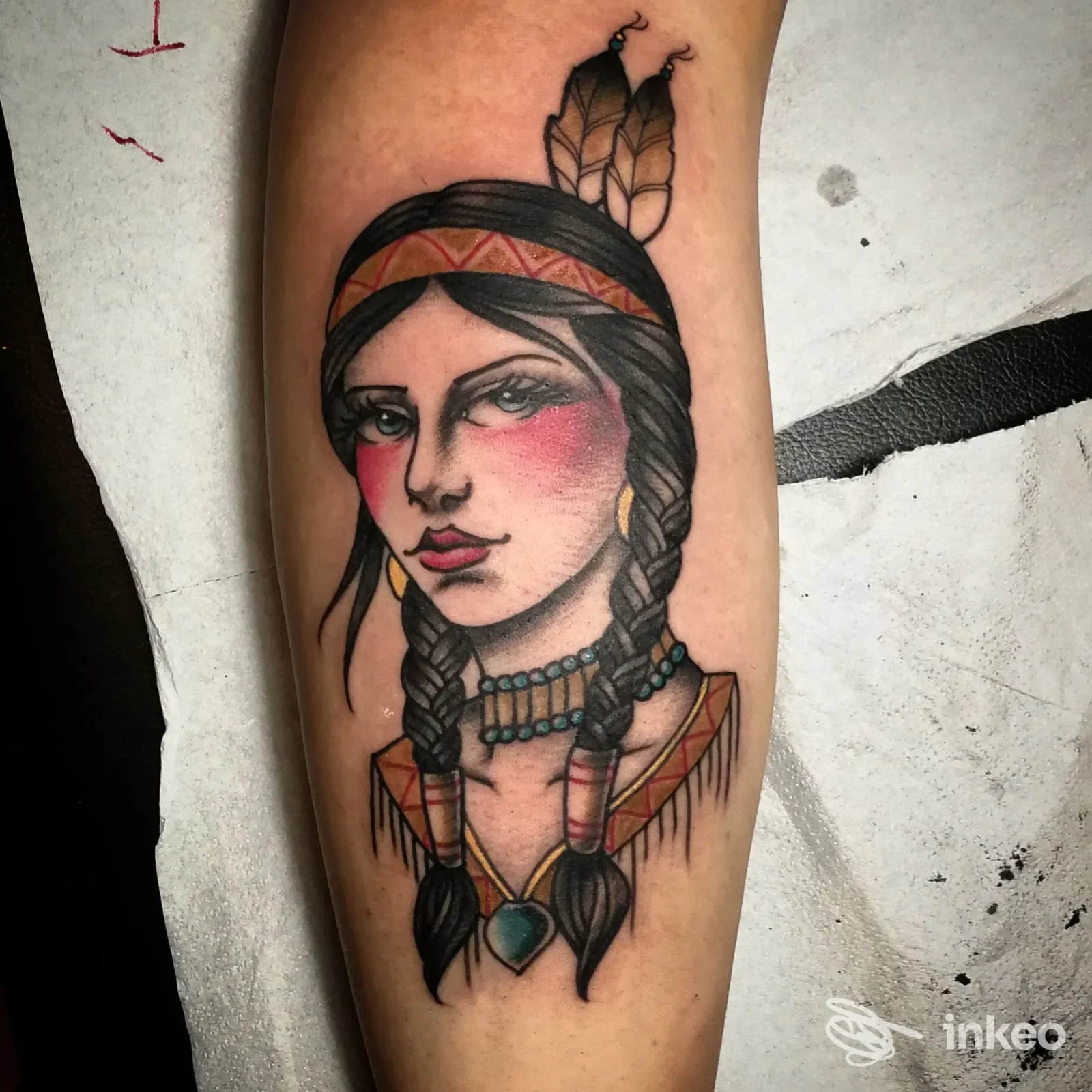 Tatuaggio su braccio raffigurante un volto femminile dallo stile nativo americano, con trecce, headband e gioielli, tonalità rosate sulle guance.