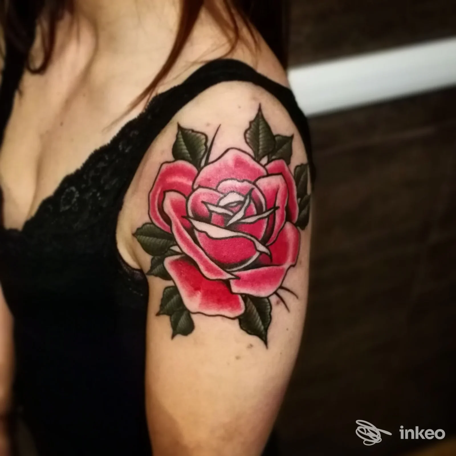 Tattoo visibile su braccio superiore: grande rosa rossa con foglie verdi, contorni neri ben definiti, senza volto visibile della persona.
