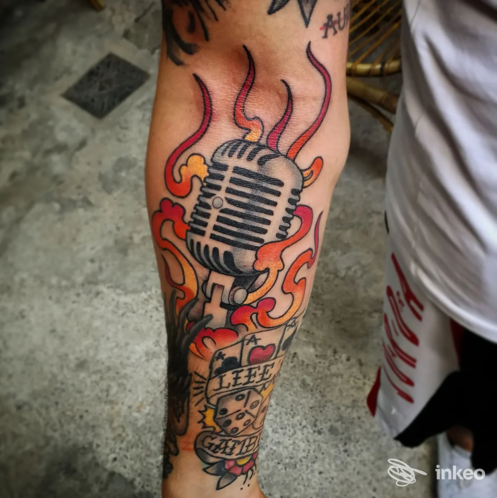 Tatuaggio sull’avambraccio raffigurante un microfono con fiamme arancioni e rosse, stile fumetto, contorni neri e colori vividi.