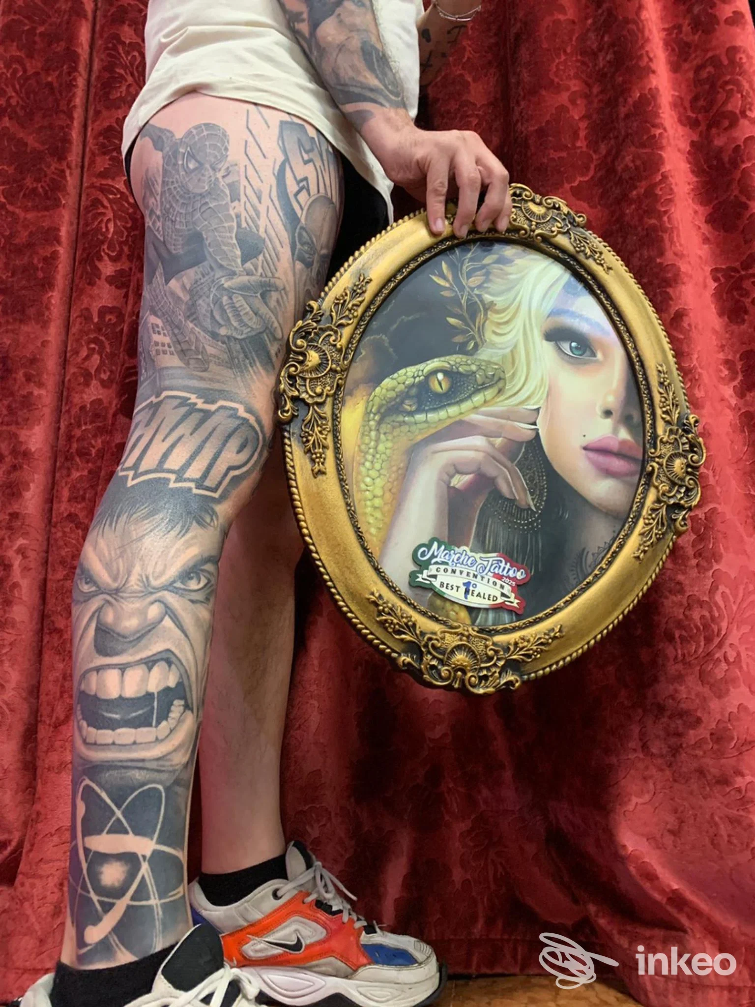 Una persona con moltissimi tatuaggi sulle gambe tiene tra le mani una cornice dorata contenente un ritratto/immagine. sfondo drappeggio rosso.