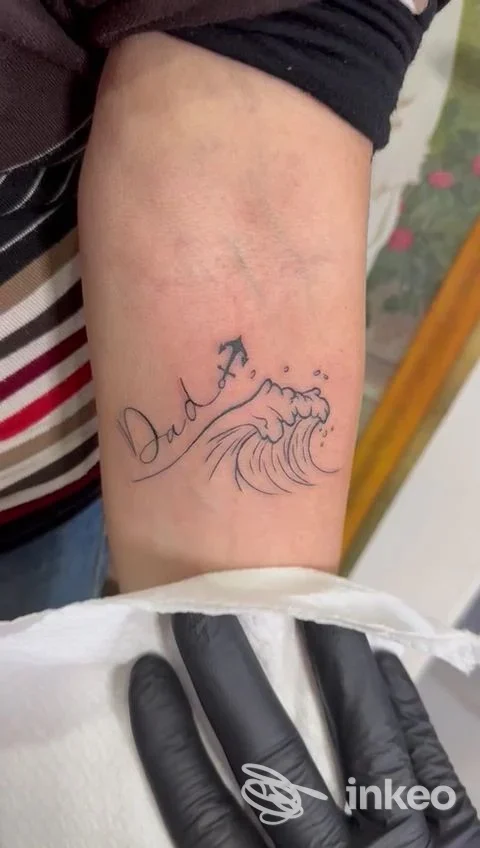 Tatuaggio sul braccio con scritta Dad e disegno di un’onda, eseguito sulla pelle verosimilmente umana.