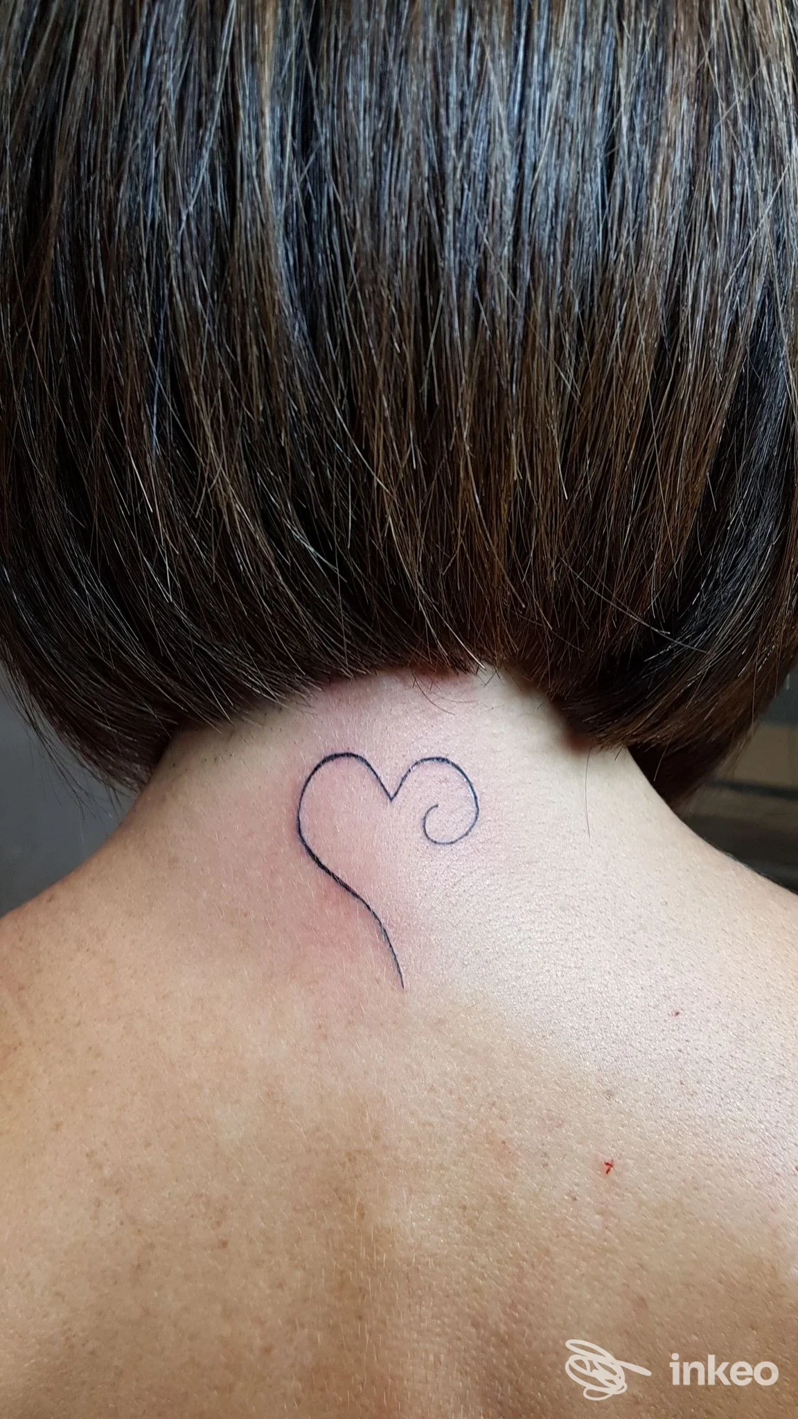 Piccolo tatuaggio a outline sul collo posteriore, forma di cuore con una piccola spirale, visibile sulla nuca, nessun volto presente.