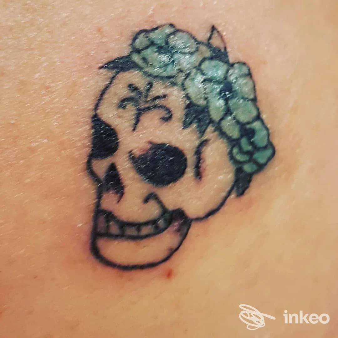 Tatuaggio su pelle di un teschio decorato con una corona di fiori verdi, visible di lato sul braccio/ascella, realizzato su pelle.