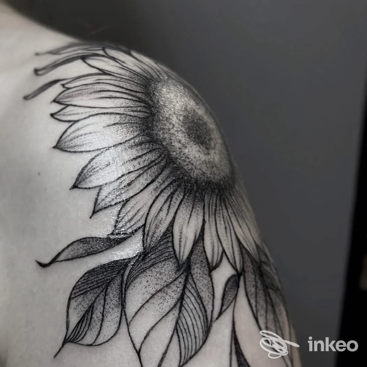 Tattoo a fioritura di girasole su una spalla, in bianco e nero, linee nette e shading, parte superiore della spalla visibile.