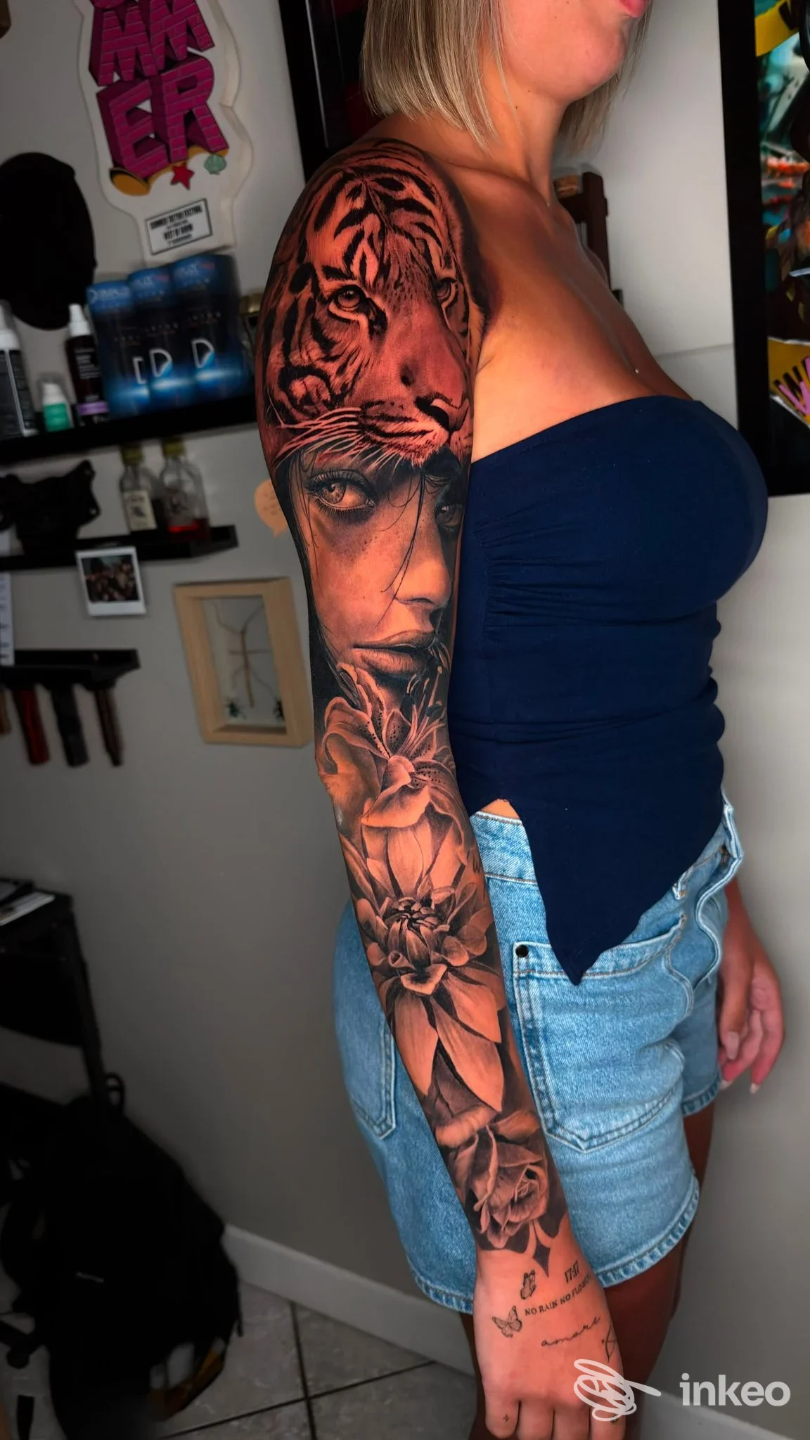 Soggetto adulto con tatuaggio a manica sul braccio sinistro, visibile di profilo laterale, indossa maglia blu, sfondo studio.