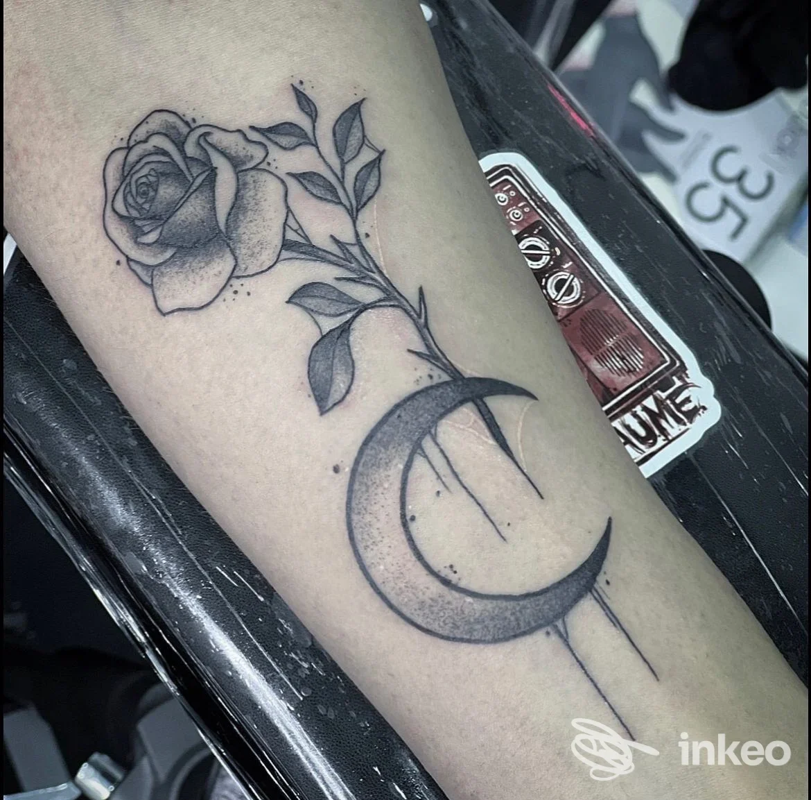 Tattoo su avambraccio: rosa stilizzata con luna crescente, in tonalità grigio-neri, disegno pulito e dettagliate foglie e linee.