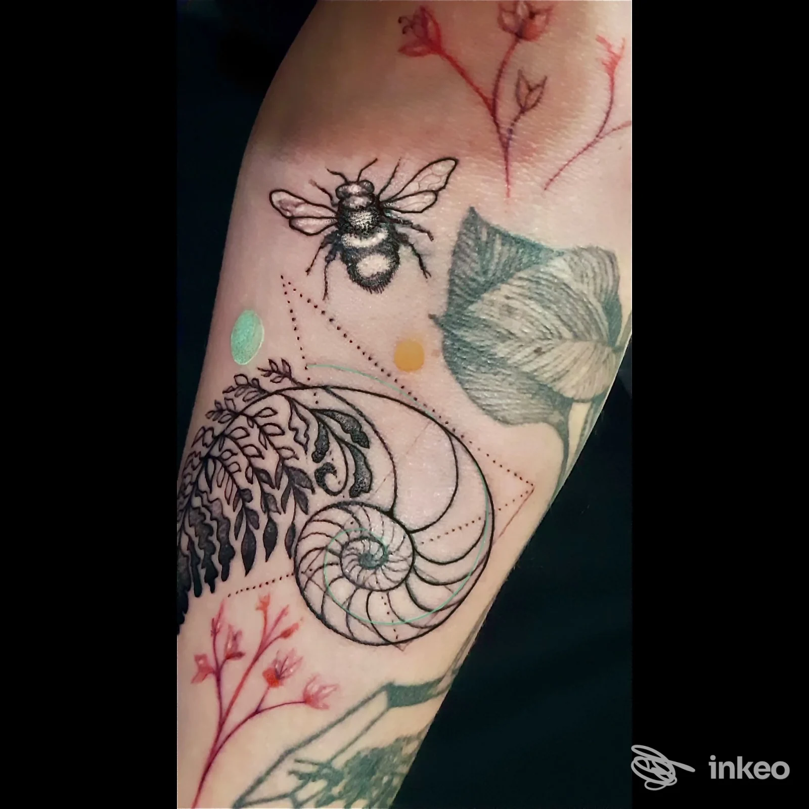 Particolare di un braccio con tatuaggi: ape nera, conchiglia ad andamento circolare e felce verde, accompagnati da motivi floreali rossi e altri elementi decorativi.