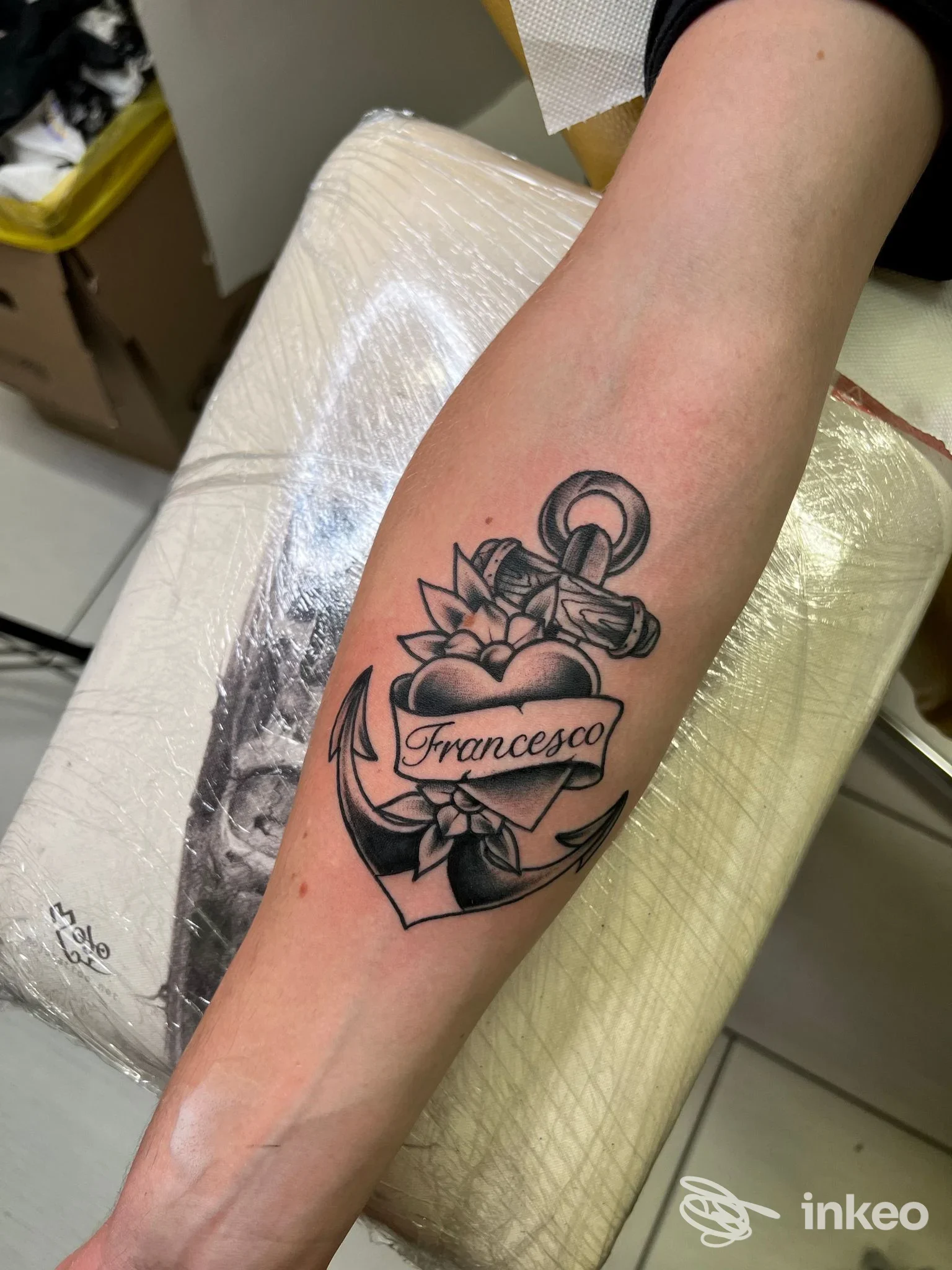 Tatuaggio visibile sull'avambraccio: ancora, cuore e fiocco intorno a un banner con il nome Francesco, realizzato con linea nera su pelle, in studio a Nocera Inferiore.