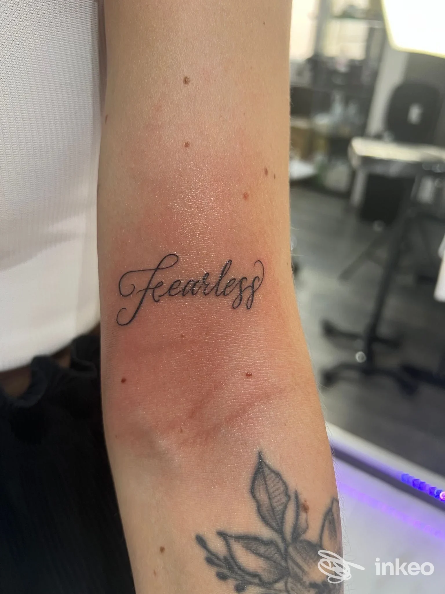 Avambraccio con tatuaggio in corsivo nero 'Fearless' visibile, accompagnato da un motivo vegetale sul polso; firma del studio Essepitattooconcept, Napoli.