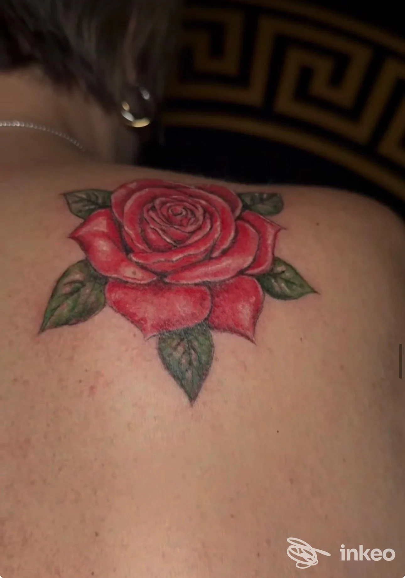 Tattoo realistico di rosa rossa su spalla, con foglie verdi, eseguito sulla pelle di una persona, visibile da dietro/di lato.