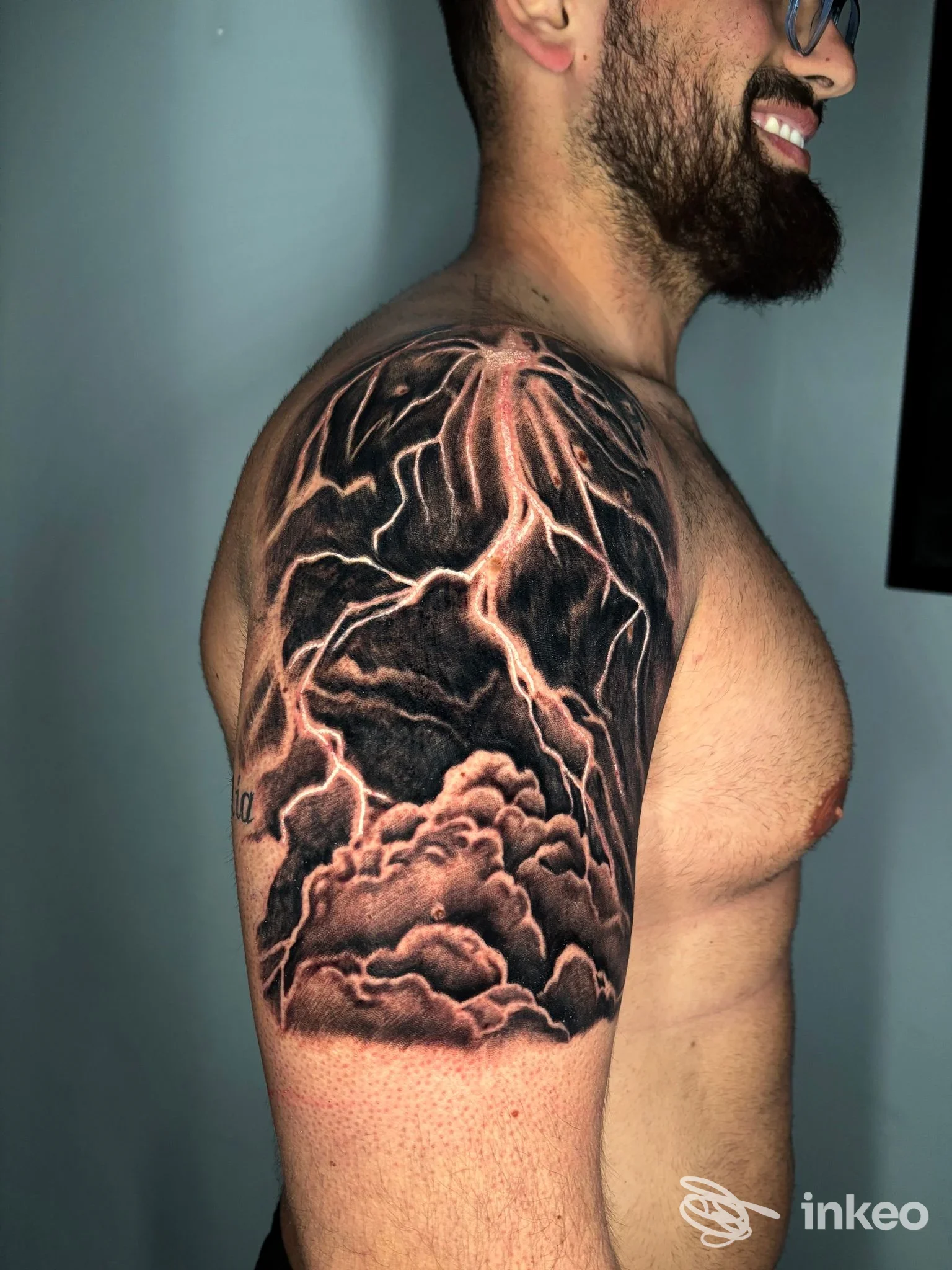 Il braccio sinistro di un uomo mostra un tatuaggio realistico di nuvole scure e fulmini rosa/rossi su sfondo nero, eseguito sulla pelle, visibile di profilo.
