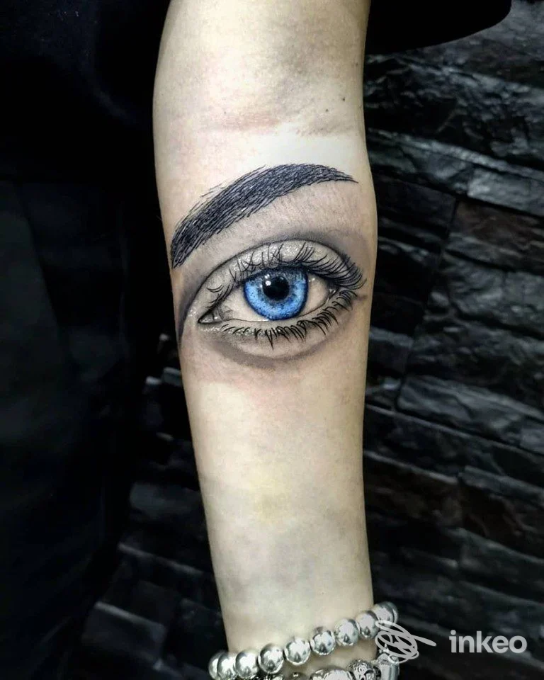 Tattoo realistico raffigurante un occhio azzurro su avambraccio, realizzato sul corpo, con sfondo scuro e bracciale in perle, studio OhMyDad Tattoo a Marano di Napoli.