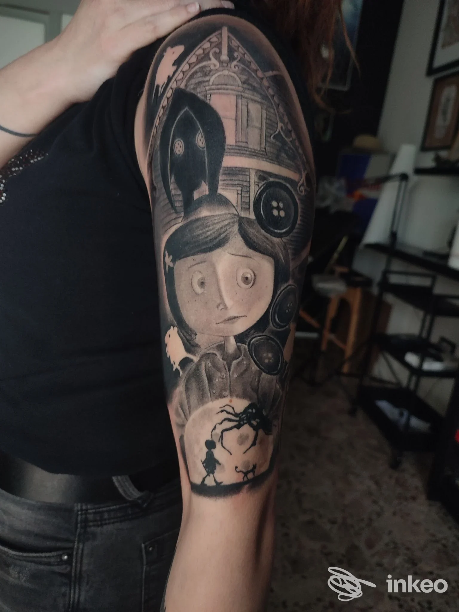 Braso di una persona con tatuaggio in bianco e nero raffigurante una figura stilizzata, con sfondo di elementi architettonici scuri e dettagli a palline, tatoo ben definito sul braccio.