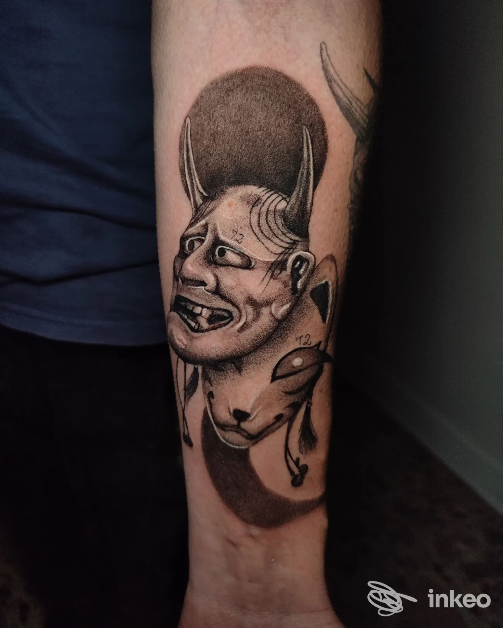 Braccio con tatuaggio a tema fantasioso raffigurante una faccia demoniaca e una creatura, eseguito in bianco e nero su pelle.