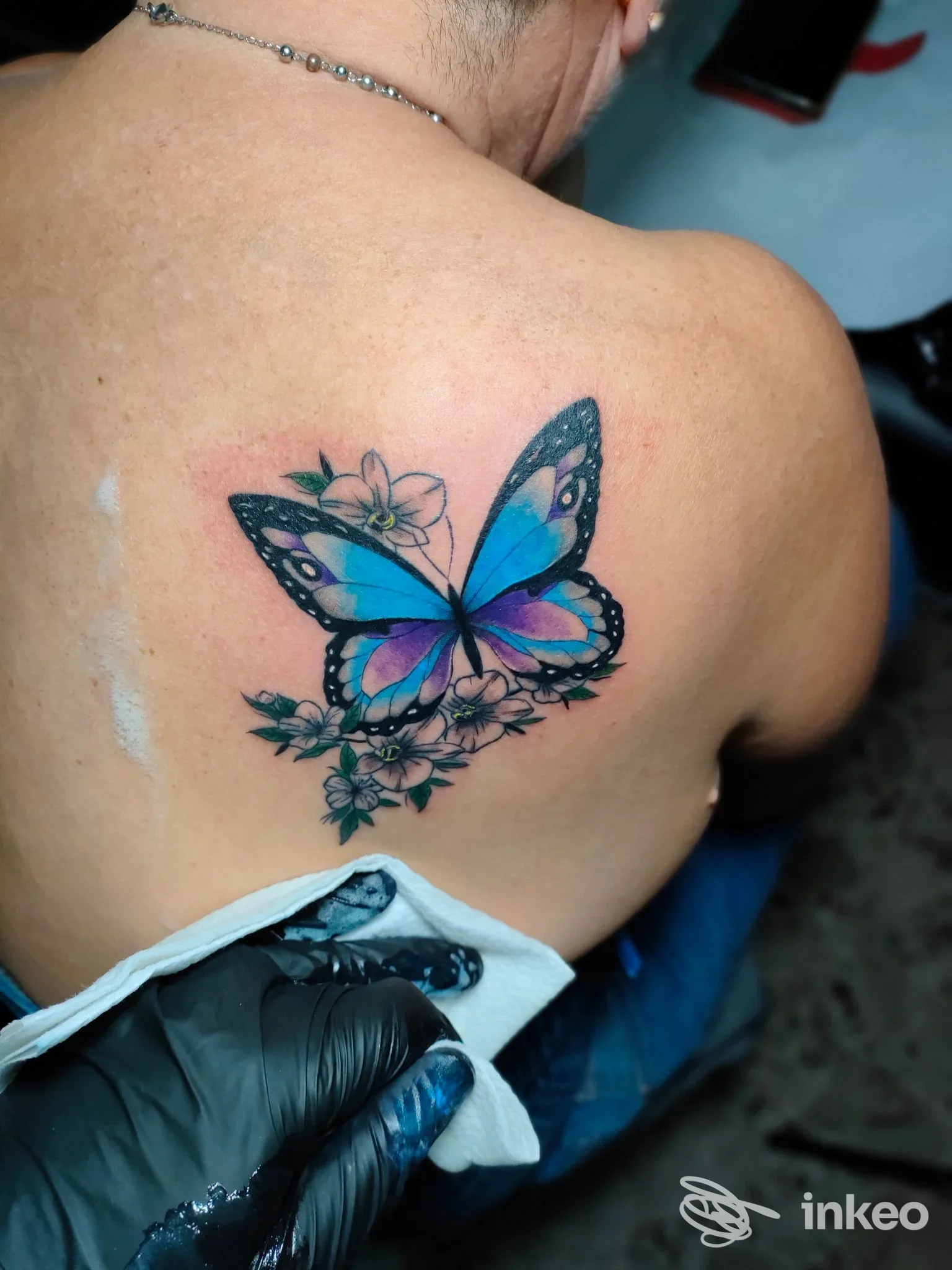 Ritocco tattile di una farfalla blu e viola sulle scapole di una persona, accompagnata da fiori; tattoo ben definito su pelle. Studio Enigma Tattoo Art Studio, Macchia.
