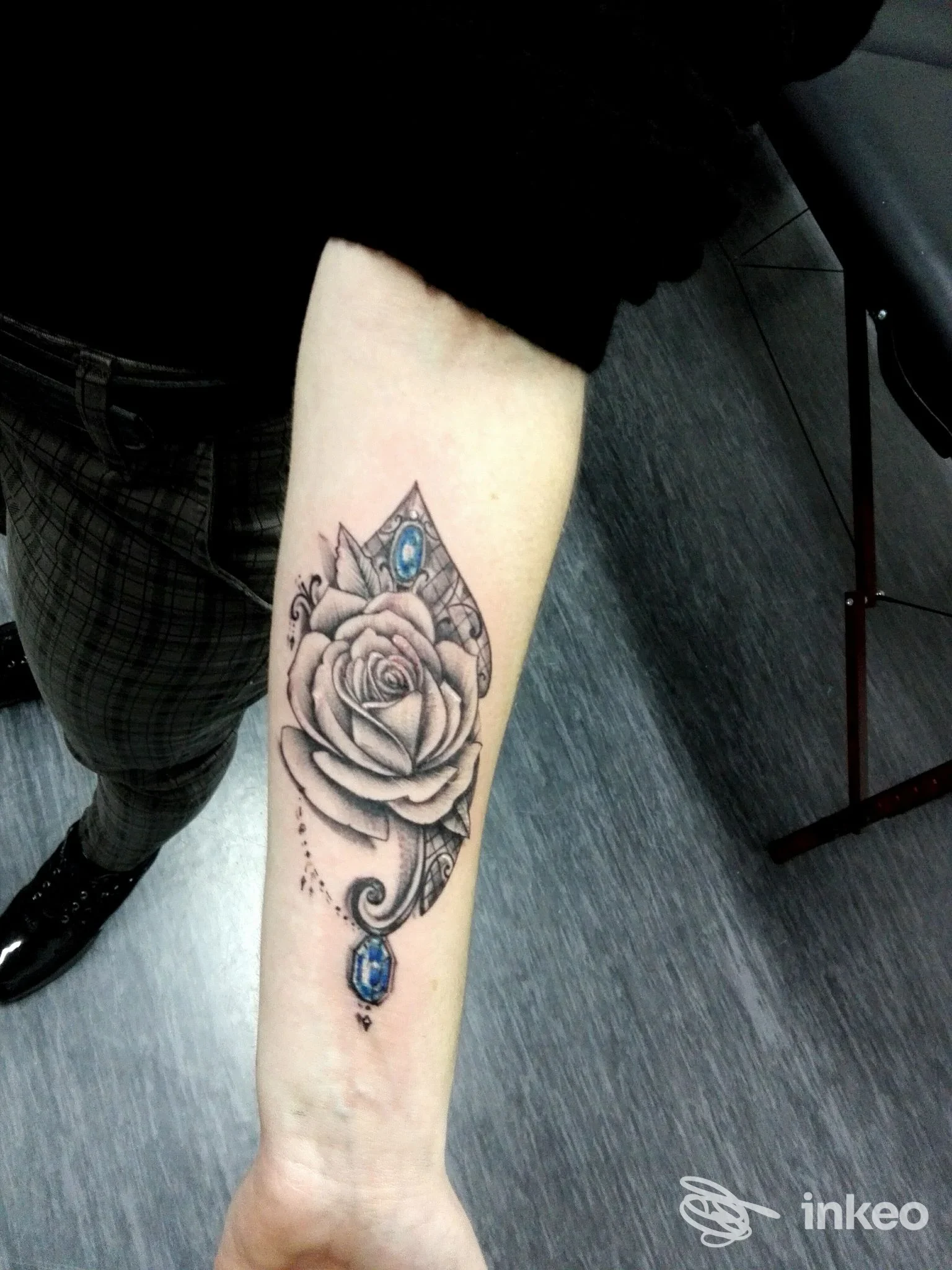 Avambraccio visibile con tatuaggio realistico di rosa, ornamenti e gemme blu. Foto di un vero tattoo su pelle, senza sovrapposizioni social o schermi.