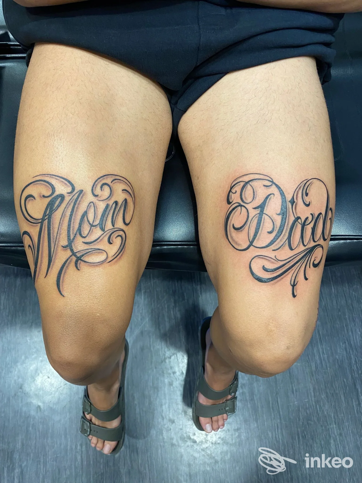 Due gambe visibili dall’alto mostrano tatuaggi in stile script su cosce, con parole Mom e Dad su pelle reale, senza volto visibile.