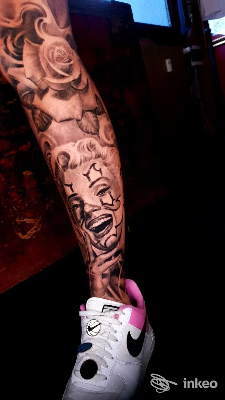 Gamba di una persona con tatuaggio completo: rosette e un volto clown stilizzato lungo il polpaccio, con una sneaker bianca, illuminazione al neon cupa.