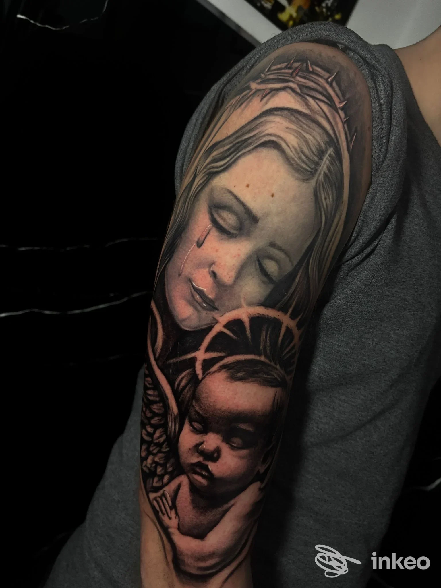 Tattoo visibile sulla spalla raffigurante una donna con pioggia di lacrime e un bambino angelo, eseguito in toni scuri e morbidi.
