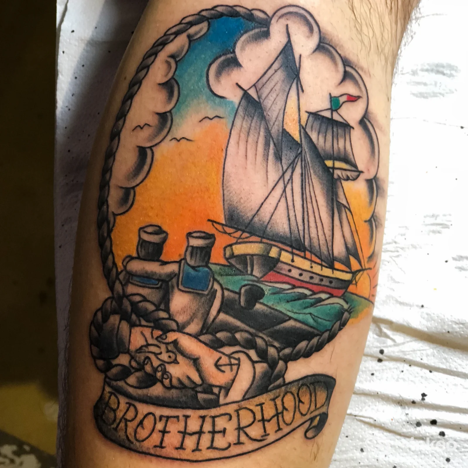 Tattoo completo sul braccio raffigurante una barca a vela ancorata con arco di corde, colori caldi al tramonto e lo stemma Brotherhood sul nastro.