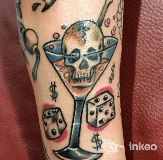 Tatuaggio visibile sulla pelle raffigurante un teschio in un bicchiere da cocktail con dadi e simboli di dollaro, colori vivaci e linee nette; Napoli, Friz Tattoo Family.