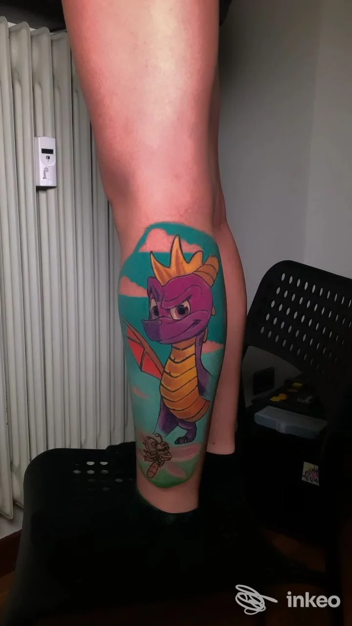 Tatuaggio colorato di un drago su polpaccio/gambe, visibile in primo piano su pelle reale, senza volti né elementi di interfaccia.
