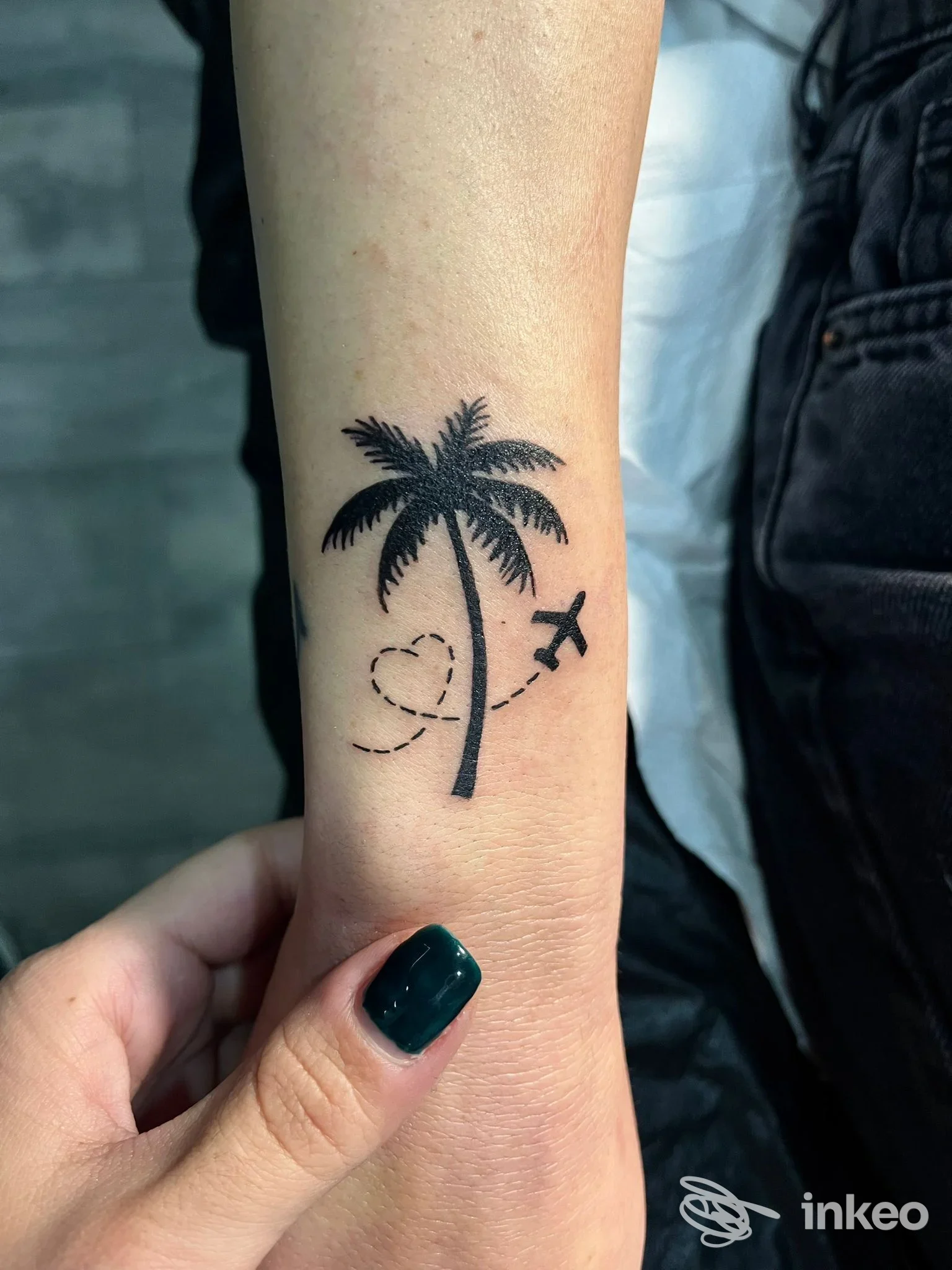 Tatuaggio su avambraccio raffigurante una palma nera con aeroplano e cuore tratteggiato, stile semplice e lineare, nessun volto visibile.
