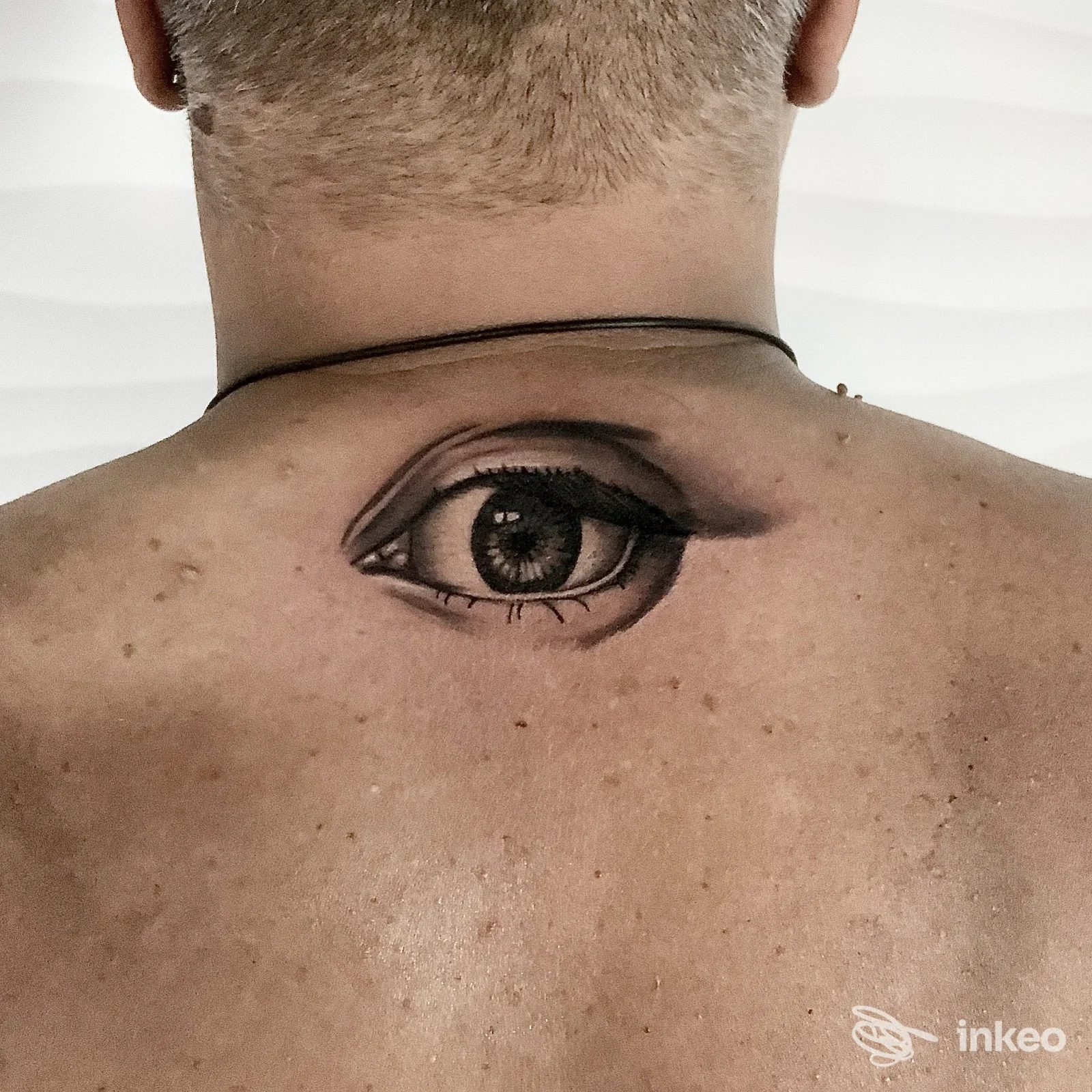 Tattoo anatomico di un occhio posizionato sulla parte alta della schiena, visibile tra collo e scapole, tatuaggio nero in stile realistico.