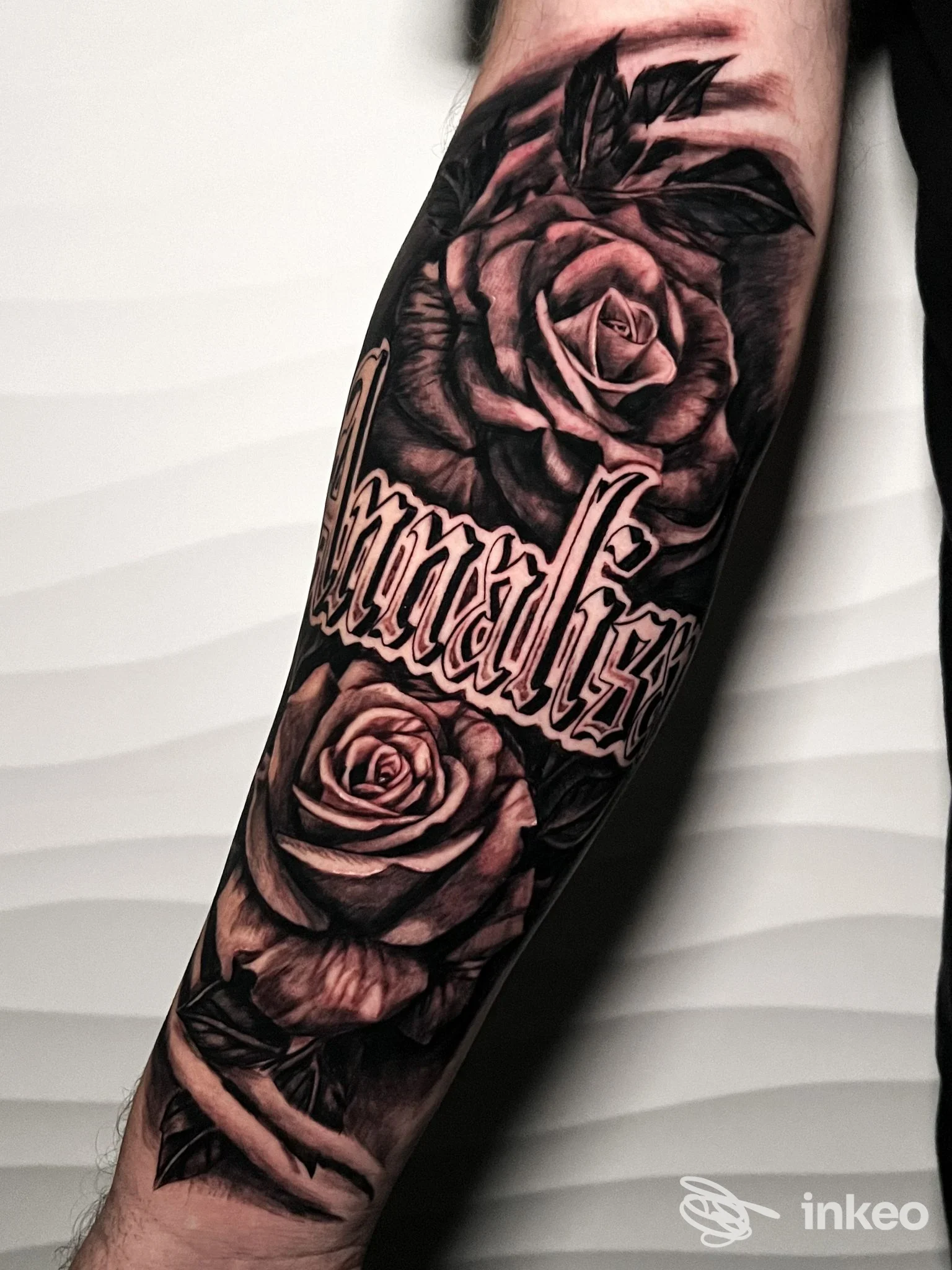Tattoo realistico in bianco e nero sull’avambraccio: due rose altamente dettagliate accompagnate da scritta ornata, realizzato sulla pelle.