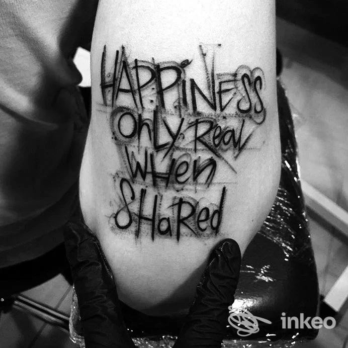 Braccio con tatuaggio testo nero: "Happiness only real when shared" scritto in stile mano libera, pelle visibile, sfondo sfocato nello studio Tattoo Brigida a Napoli.