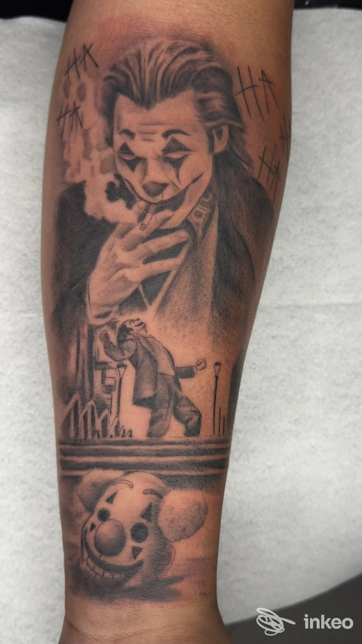 Tatuaggio in bianco e nero su avambraccio raffigurante il Joker che fuma, con figure e barre decorative sullo sfondo; realizzato dallo studio Take Your Tattoo, Battipaglia.