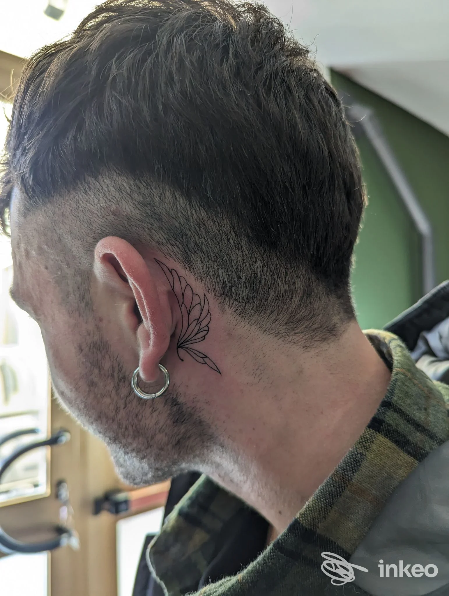 Vista laterale del collo di una persona, mostra un tatuaggio floreale dietro l'orecchio, con capelli corti e orecchino tondo; illuminazione naturale in studio a Le Officine Tattoo Studio, Laveno-Mombello.