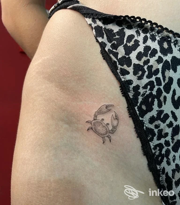 Piccolo tatuaggio a tema granchio sulla pelle della spalla di una persona, con linee sottili e tonalità nere/grigie.