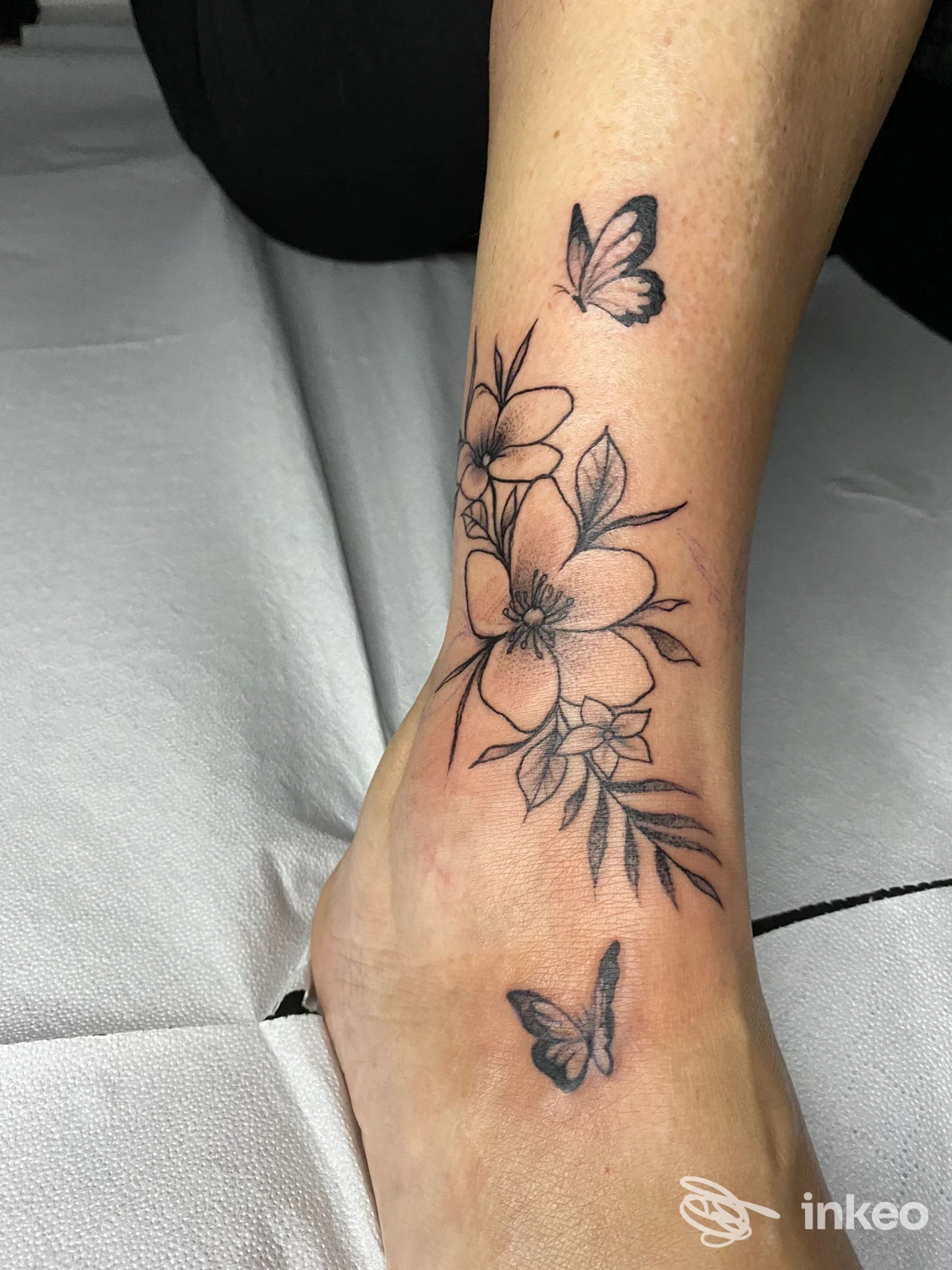 Tattoo in bianco e nero di fiori e farfalle sull’osso della caviglia/tallonare, realizzato direttamente sulla pelle, stile lineare e delicato.
