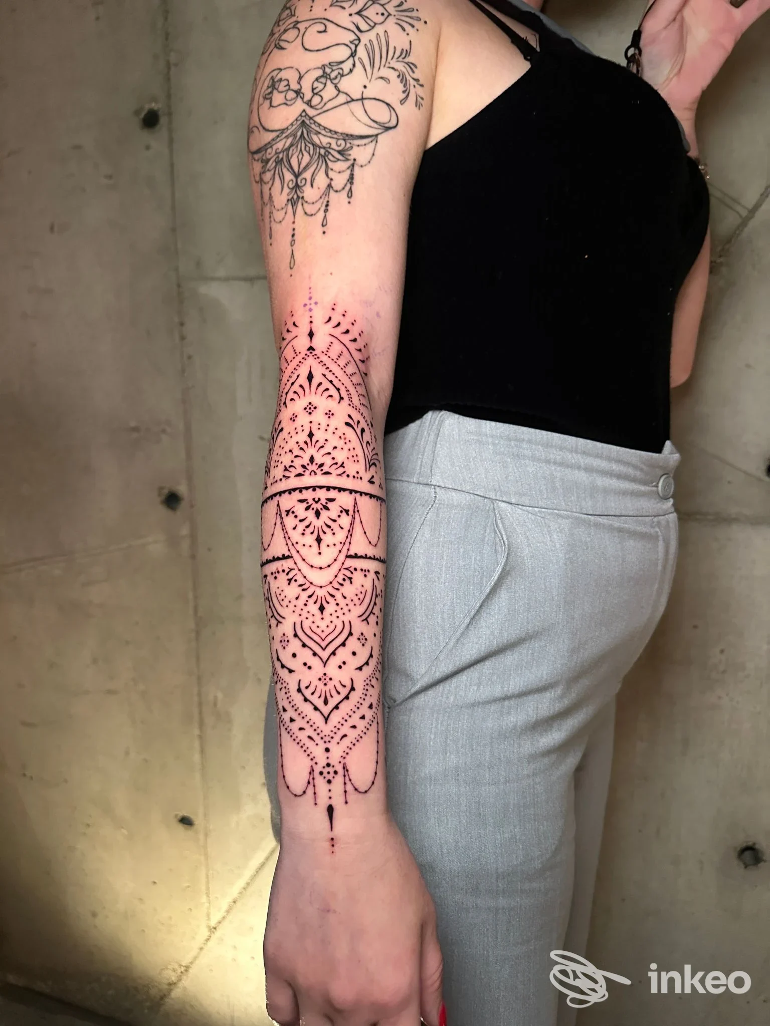 Particolare di un braccio con tatuaggio a motivi geometrici e puntinati in bianco e nero, su pelle, con sfondo muro in cemento; persona indossa top nero.