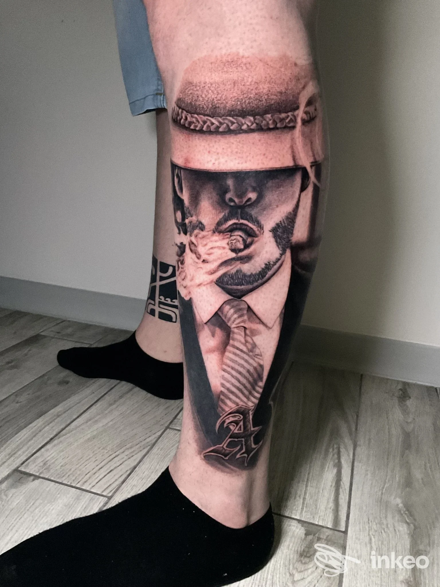 Tattoo realizzato sulla gamba, ritraente una figura in abito e cappello, in stile realistico, con tonalità in bianco e nero; visibile l’aria di un cappuccio o ombra sul volto.
