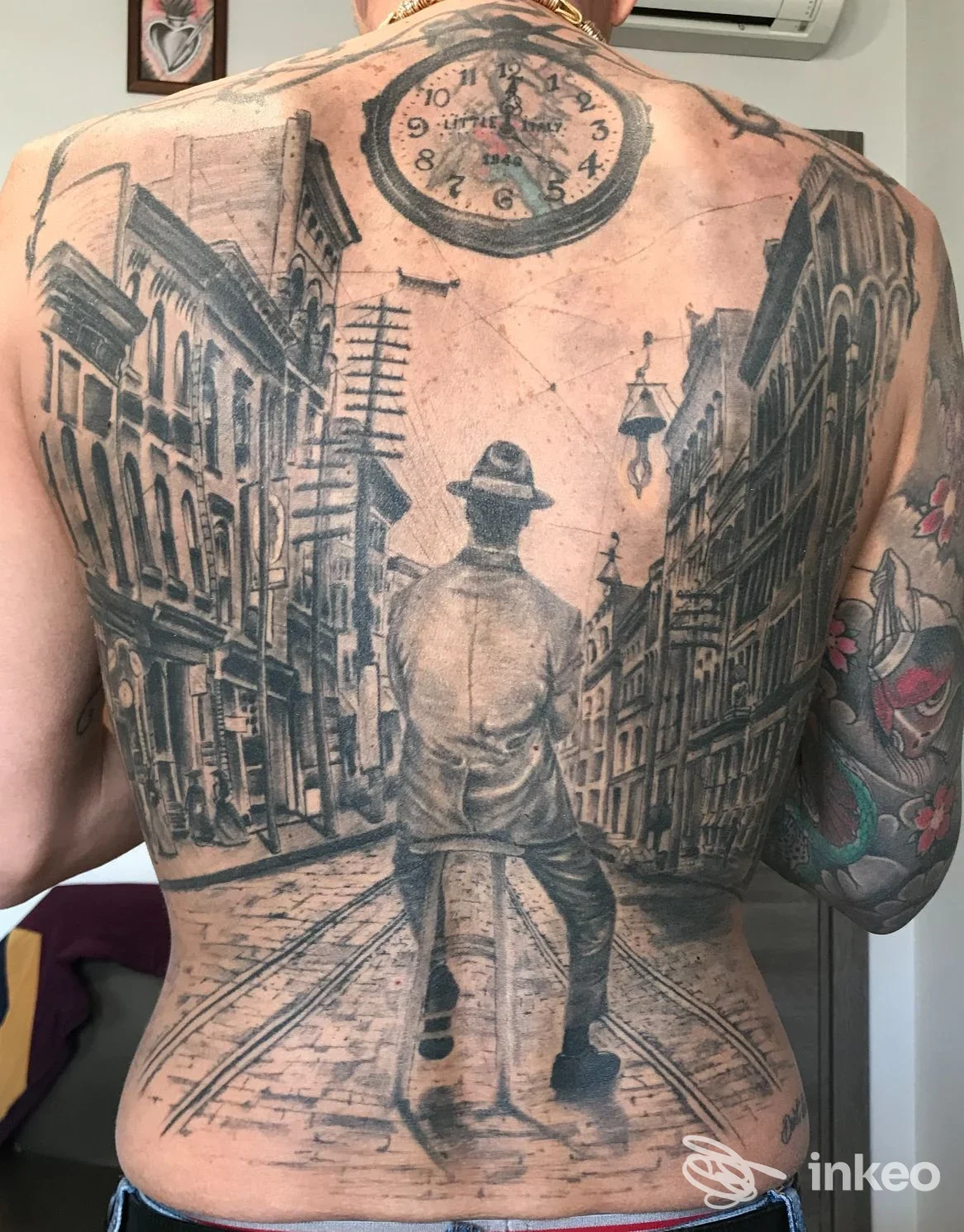 Grande tatuaggio sul dorso che raffigura una scena urbana con una figura al centro e un grande orologio circolare in alto; stile in bianco e nero su pelle chiara; studio Old River Ink, Aversa.