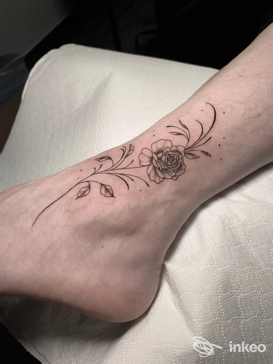 Tattoo floreale nero disegnato sulla pelle della caviglia/piede, con una rosa centrale e decorazioni lunghe e sottili