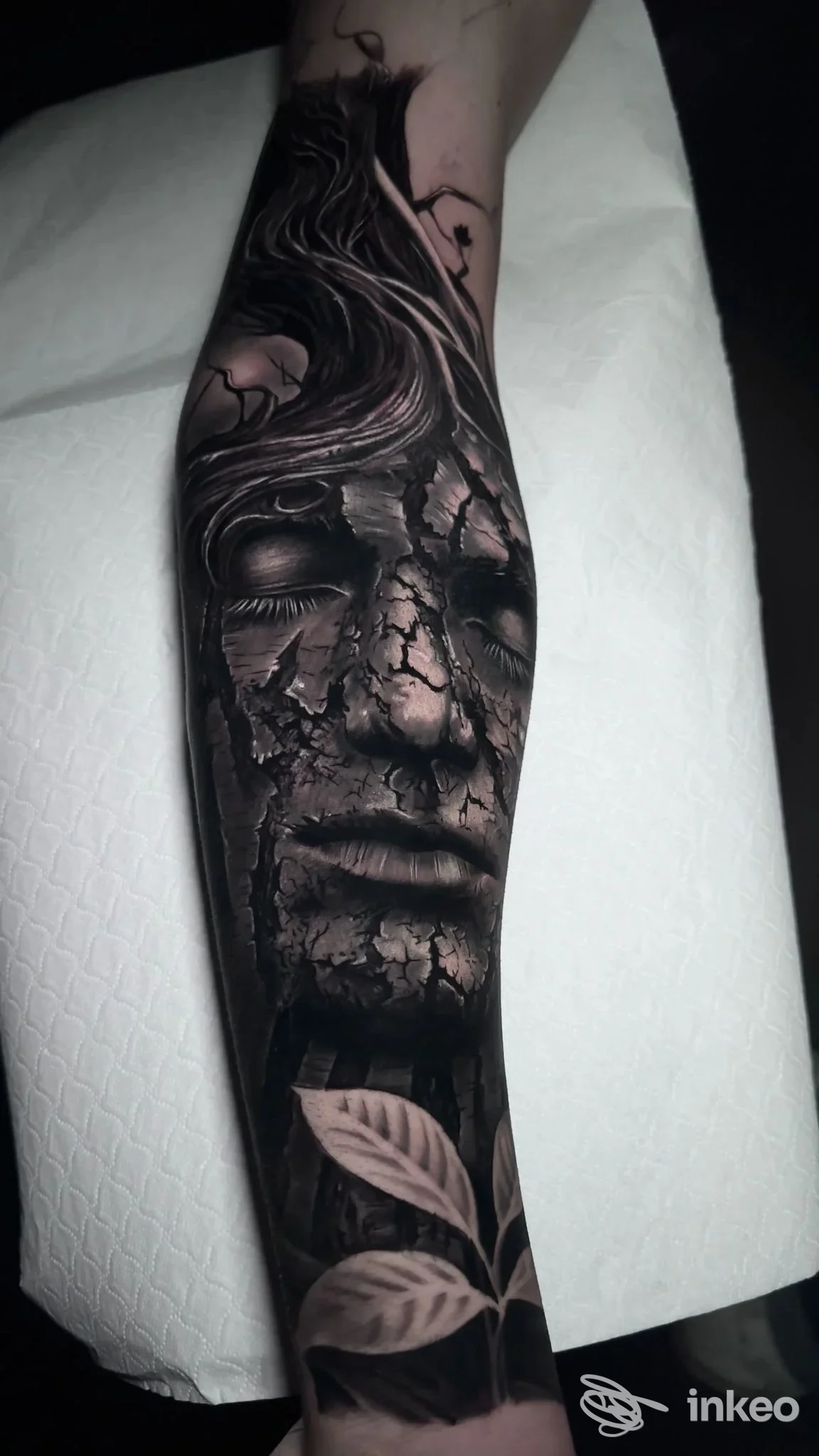 Avambraccio sinistro con tatuaggio realistico in bianco e nero: volto aperto in stile scultura e rami e foglie nella parte inferiore, alto livello di dettaglio e contrasti, illuminazione singola.