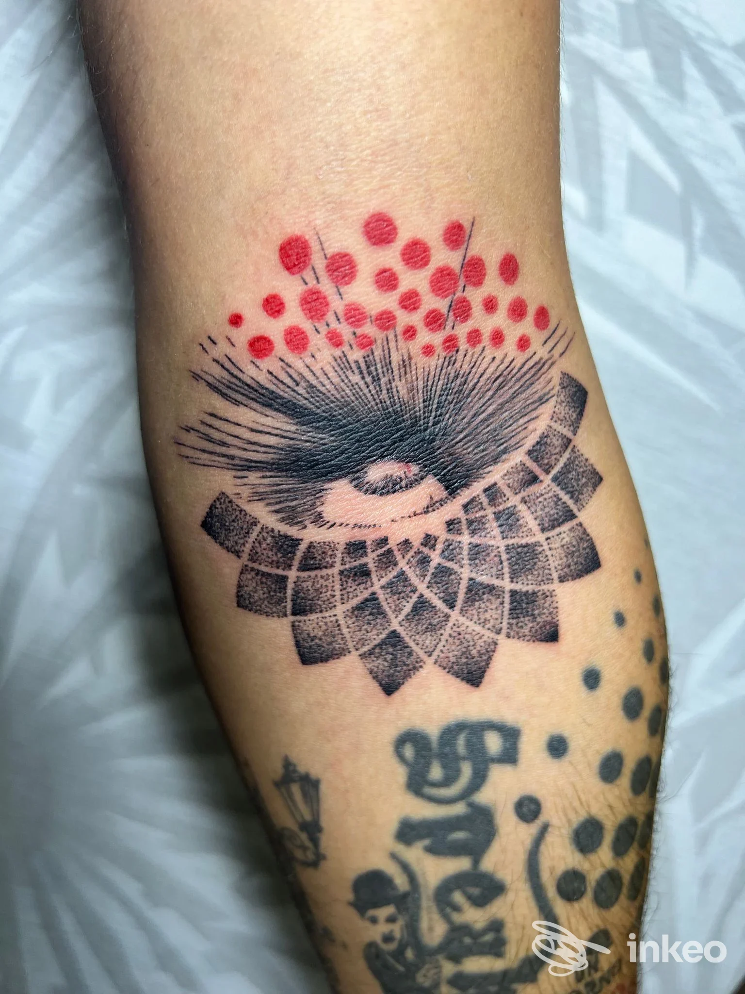 Disegno di tatuaggio sull'avambraccio raffigurante un occhio stilizzato con ciglia e motivi geometrici a formare una specie di mandala, con puntini rossi sopra.