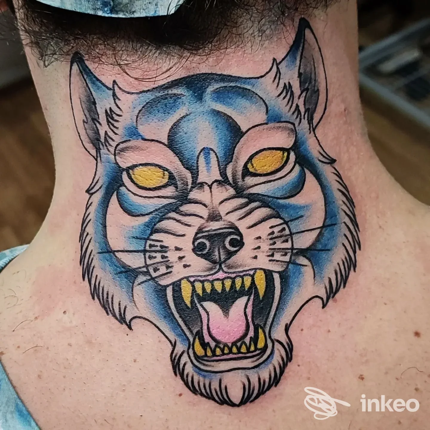Collo tatuato con la testa di un lupo blu, con baffi neri, occhi gialli e fauci aperte, realizzato come tatuaggio sul retro del collo, colori vividi e linee nette.