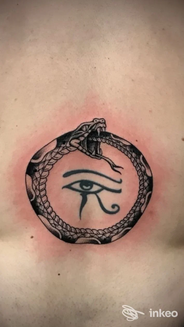 Tatuaggio circolare con serpente che morde la coda avvolto attorno a un occhio di Horus, eseguito sulla pelle, con contorni nitidi e leggero rossore intorno.