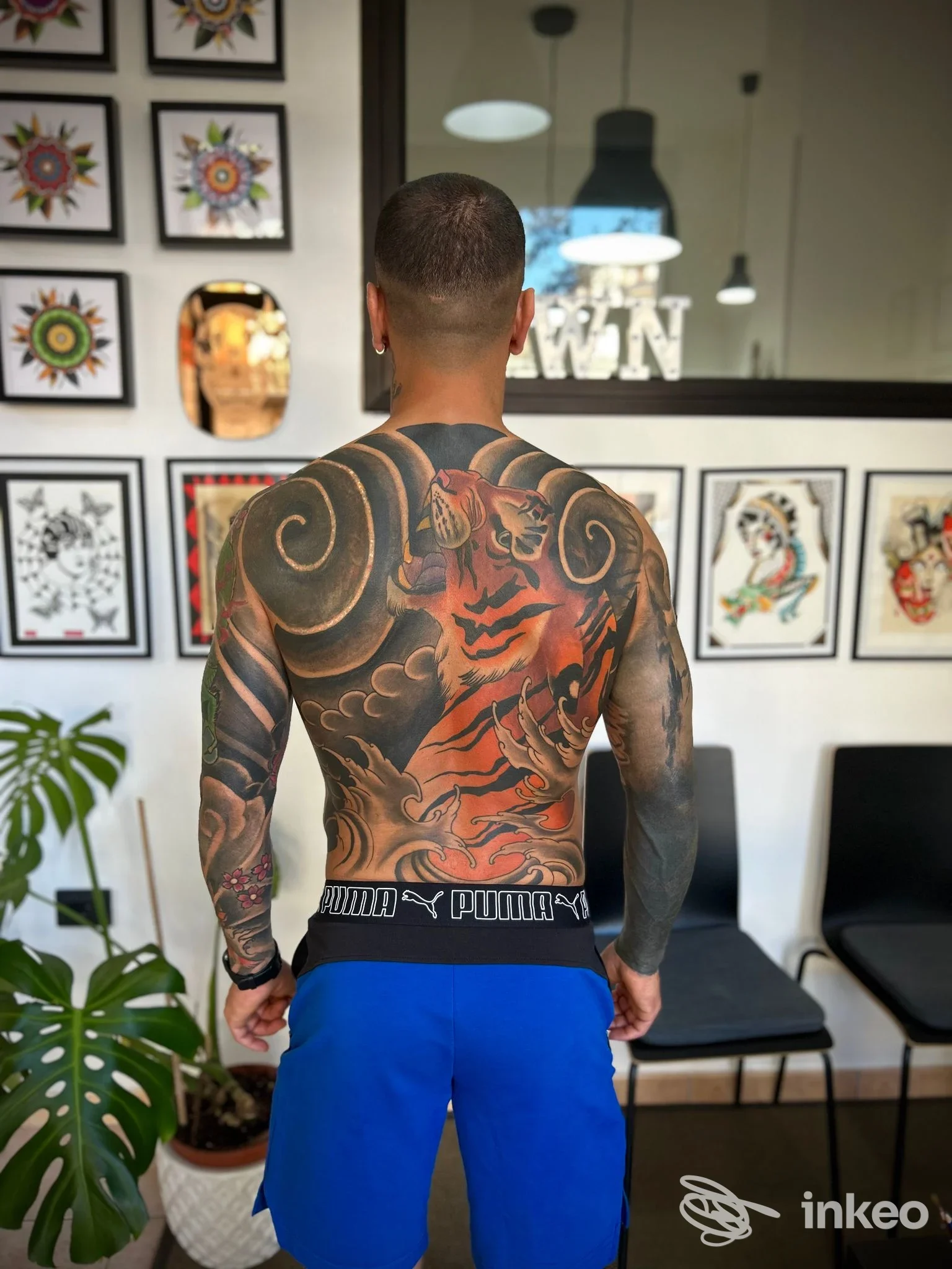 Tatuaggio completo sulla schiena di un uomo, realizzato da White Noise Tattoo Lab a Eboli, con figure in toni neri, grigi e arancioni; shorts blu e sfondo di tattoo gallery.