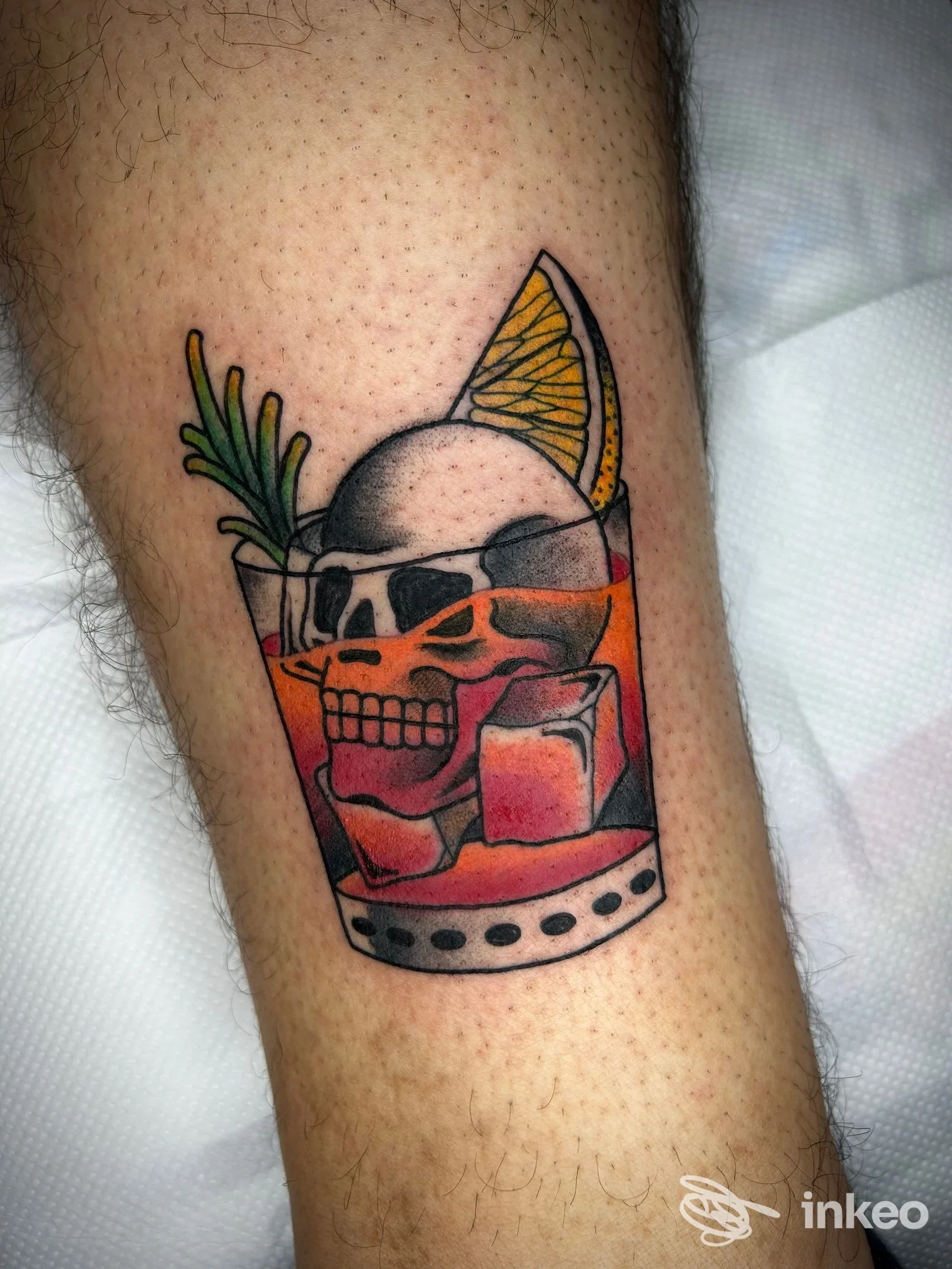 Tatuaggio a colori su pelle raffigurante un bicchiere con teschio, ghiaccio e fetta d'arancia, con un rametto verde e riflessi. studio White Noise Tattoo Lab, Eboli.