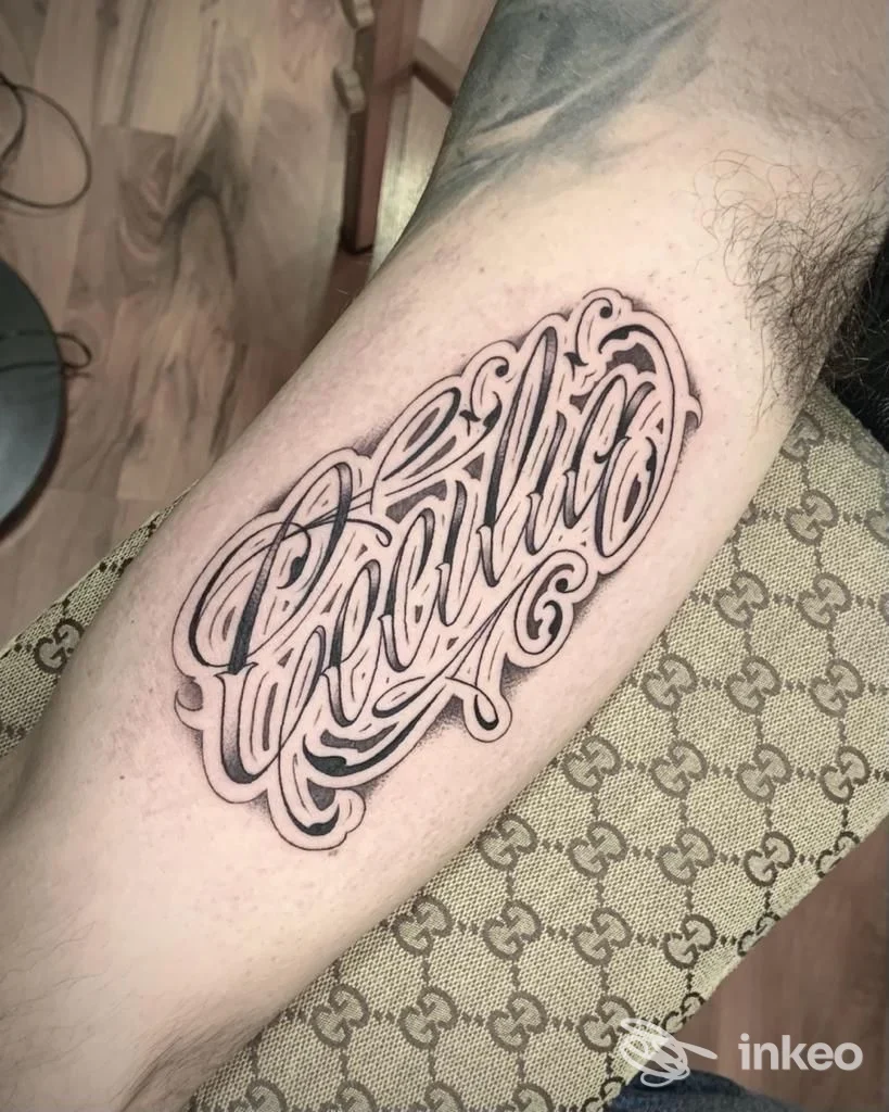 Tattoo lettering elaborato sul braccio, stile calligrafia ornata in nero su pelle chiara