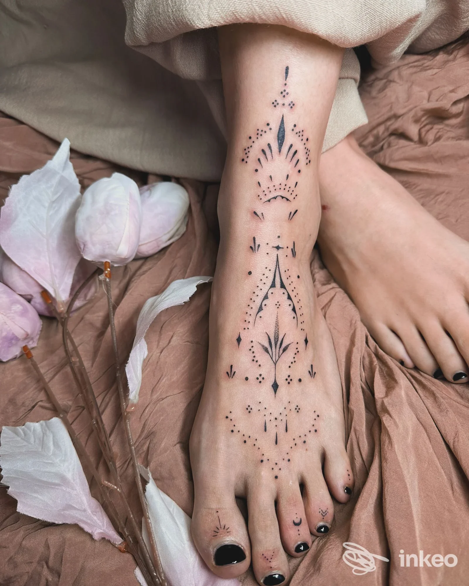Piede e parte bassa della gamba di una persona, tatuaggio geometrico a puntini lungo il dorso del piede, con dettagli decorativi, su sfondo di tessuto beige.