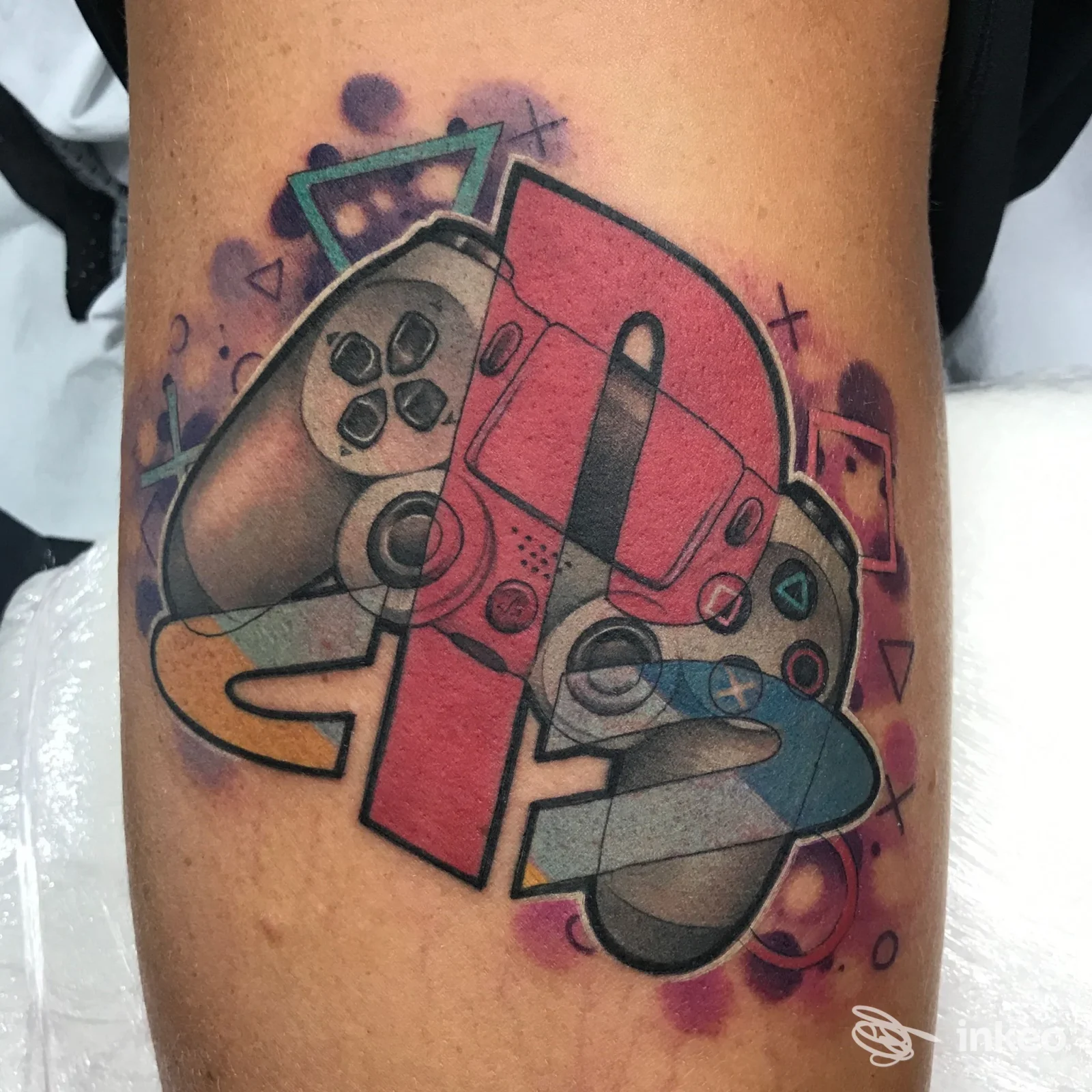 Tattoo a colori su pelle raffigurante un controller di videogiochi stilizzato, con sfondo viola e dettagli geometrici.