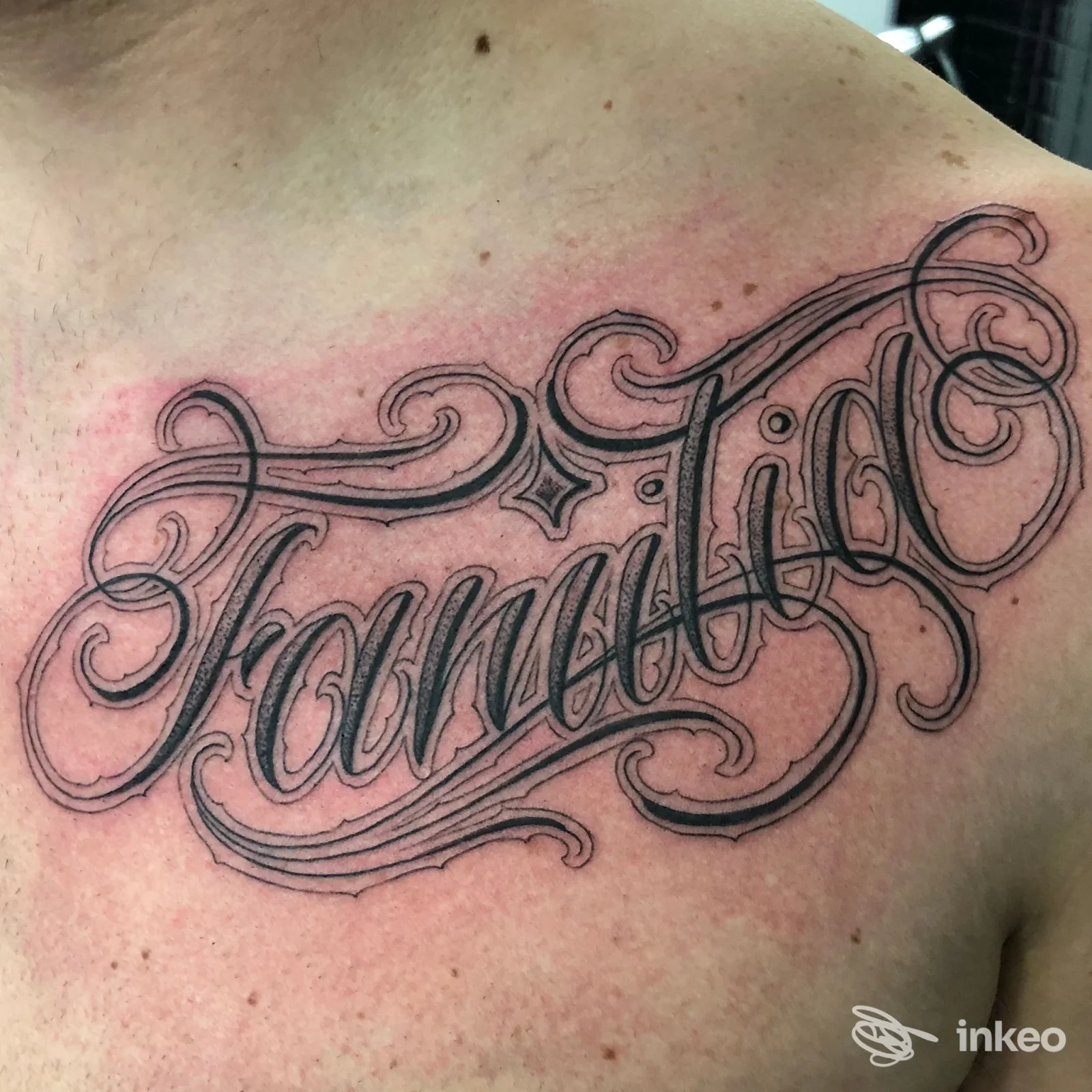 Tattoo sulla pelle con una scritta ornamentale in stile script, sembra leggere "Familia" lungo la spalla; linee nere finemente dettagliate.