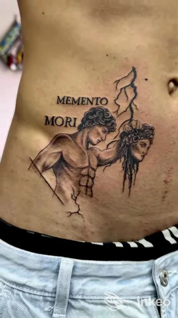 Tatuaggio realistico su pelle sul fianco, raffigurante due figure con scritte MEMENTO MORI, visibile su torace/addome; tonalità scure e linee nette.