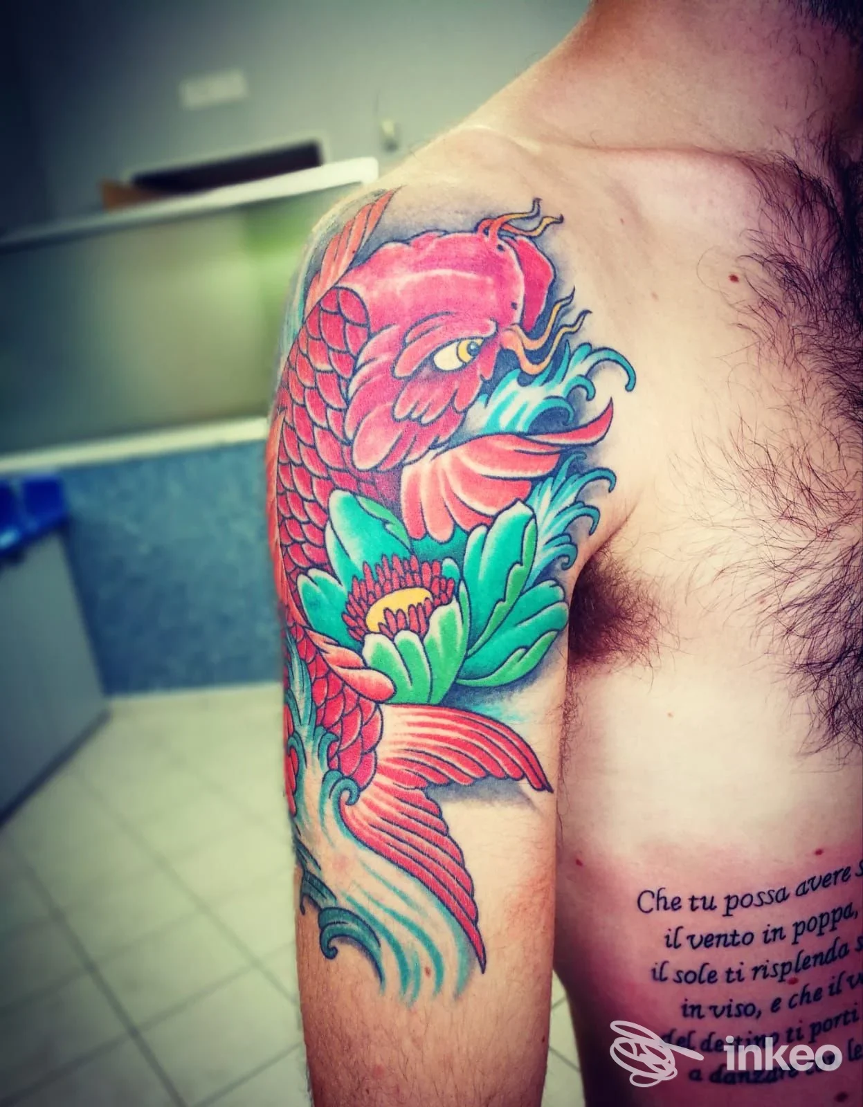 Una spalla di uomo con tatuaggio colorato di koi rosso e fiore verde, visibile su braccio e spalla.